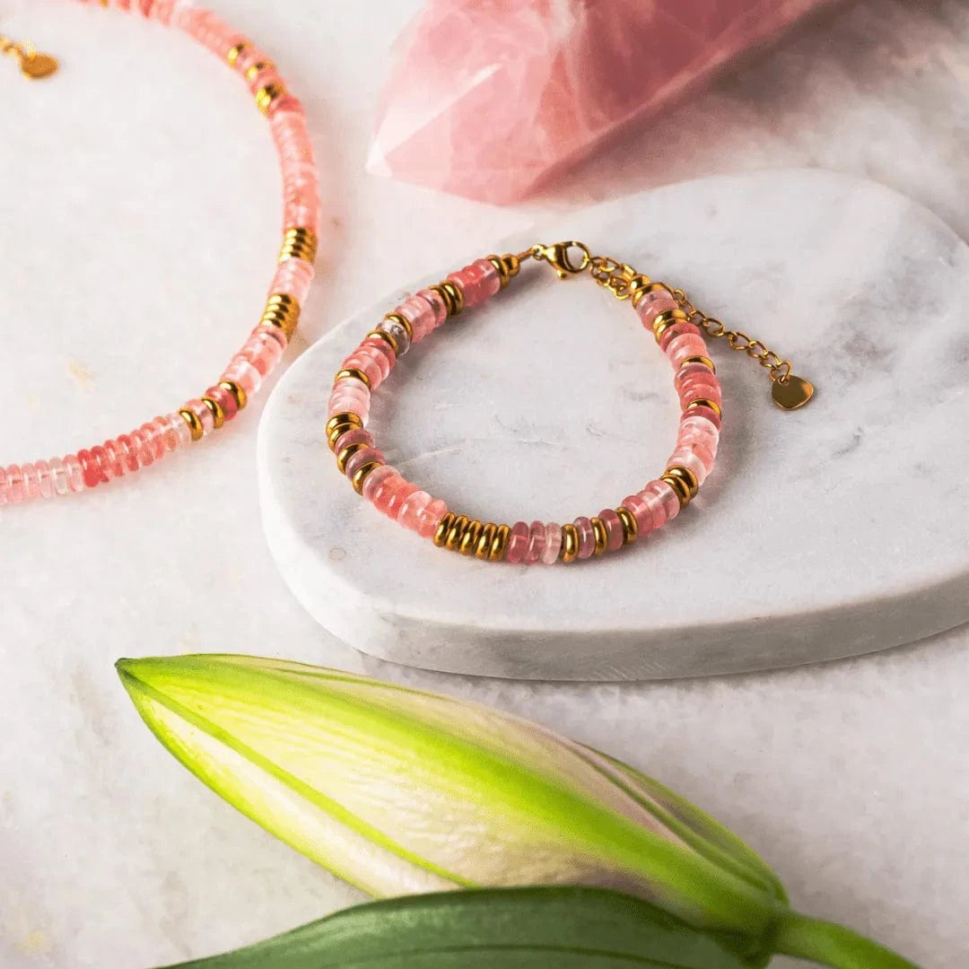 Pink beaded bracelet with gold accents on a marble surface with a pink crystal and green leaf.