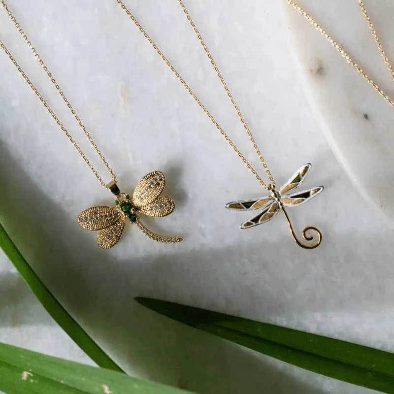 Two gold dragonfly necklaces on a marble surface with green leaves.