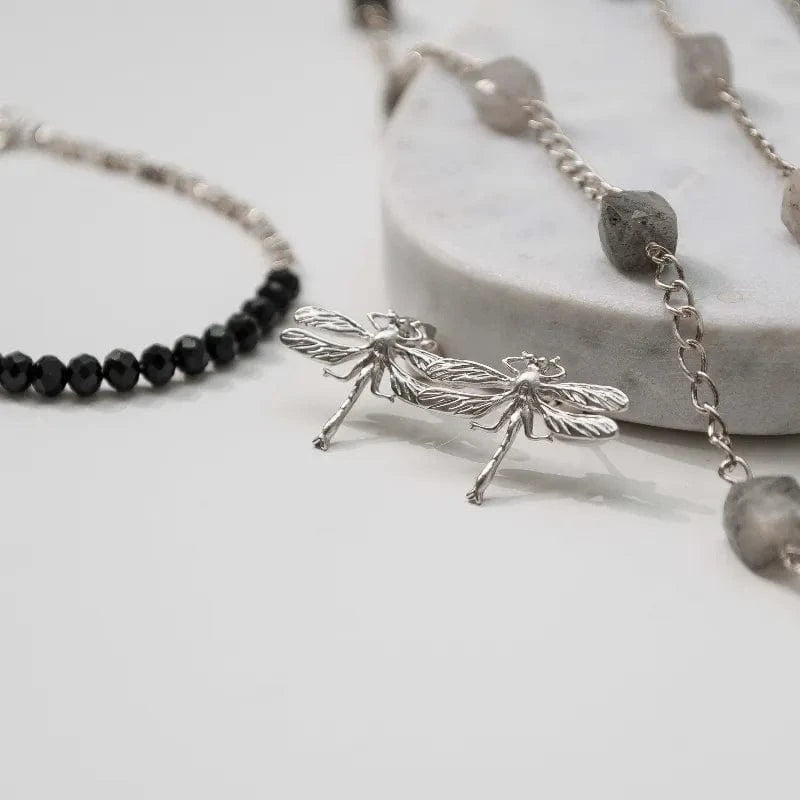 Silver dragonfly stud earrings and black beaded bracelet on a light gray surface