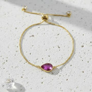Gold bracelet with a purple teardrop-shaped gemstone on a textured white surface