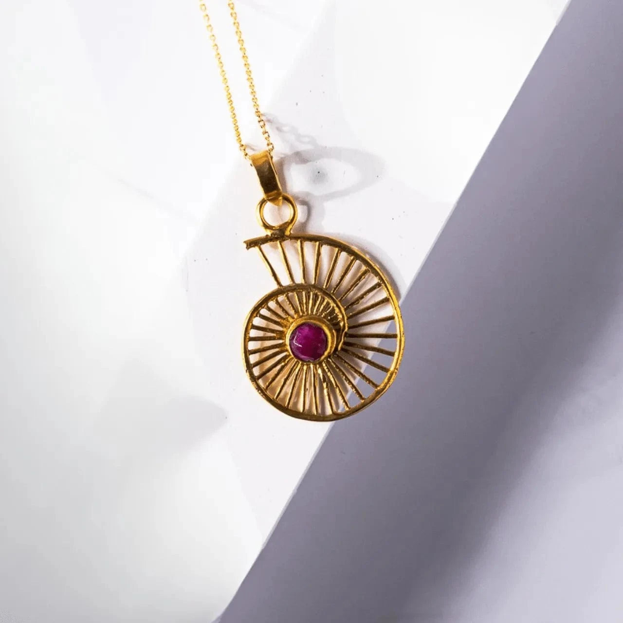 Gold necklace with a circular pendant featuring a red gemstone on a white background
