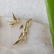Gold bird and leaf earrings on a white stone surface with a green leaf in the background