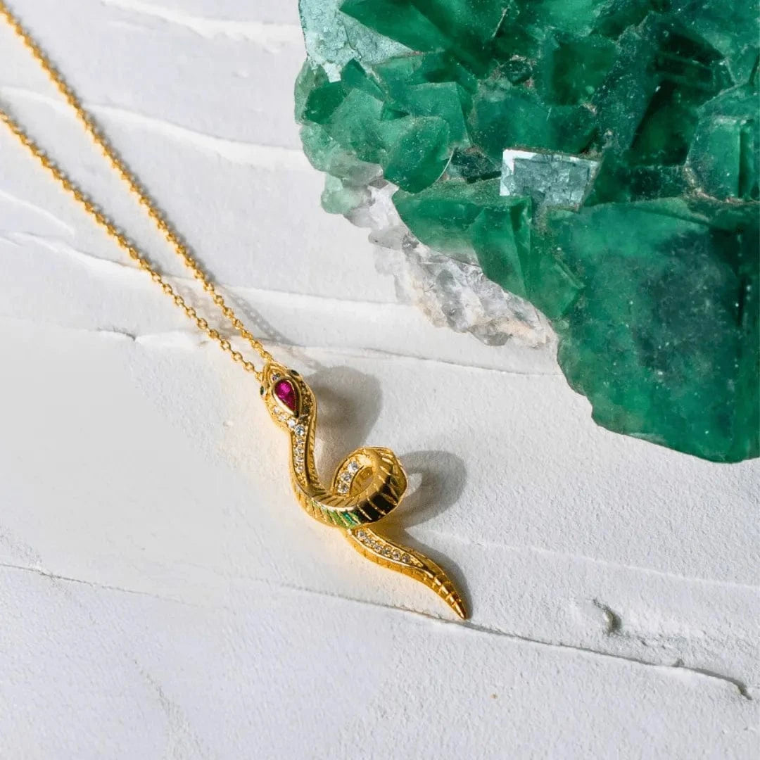 Gold snake-shaped pendant with gemstones on a white surface with green crystals in the background