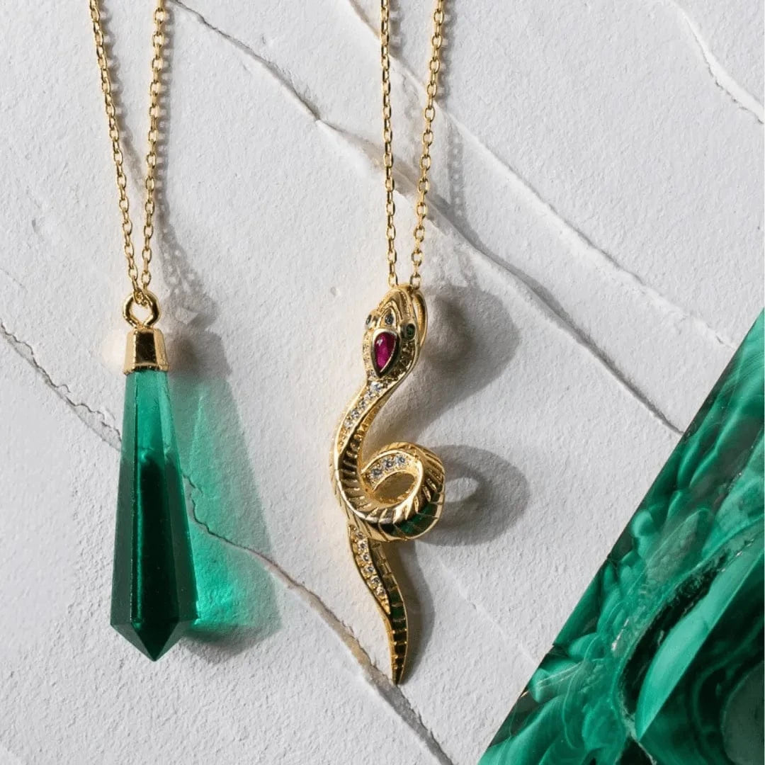 Two gold necklaces with green and red gemstone pendants on a light marble background.