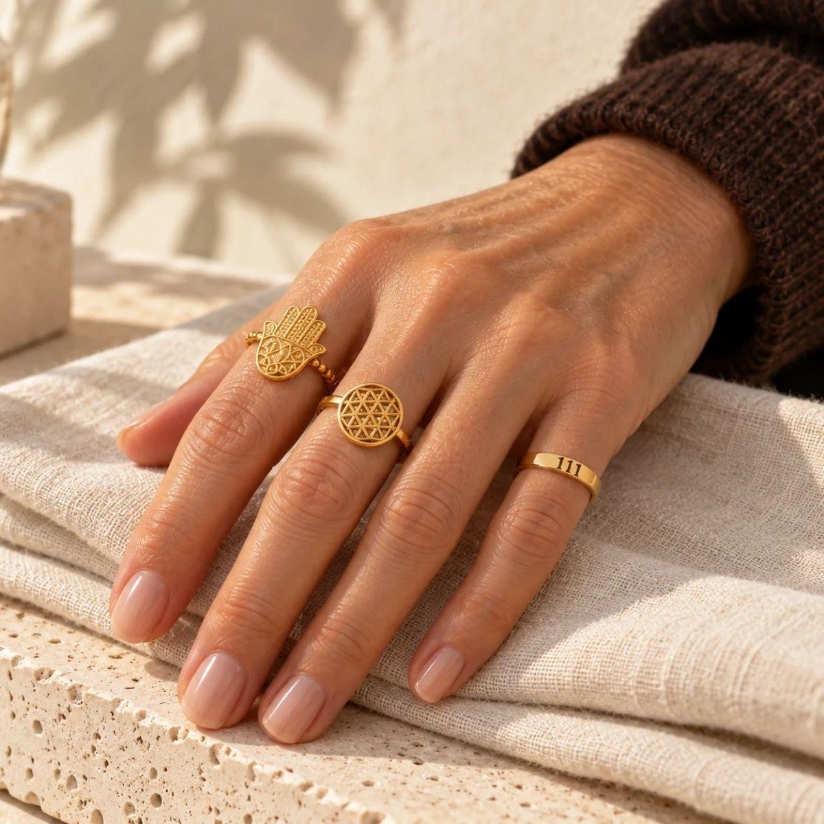 Hand wearing a gold hamsa ring, flower of life ring and angel number ring — what a ring on each finger means