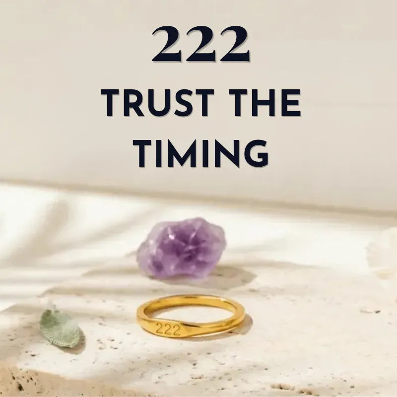 Gold ring with engraved numbers on a stone surface with crystals, accompanied by text '222 Trust the Timing'.