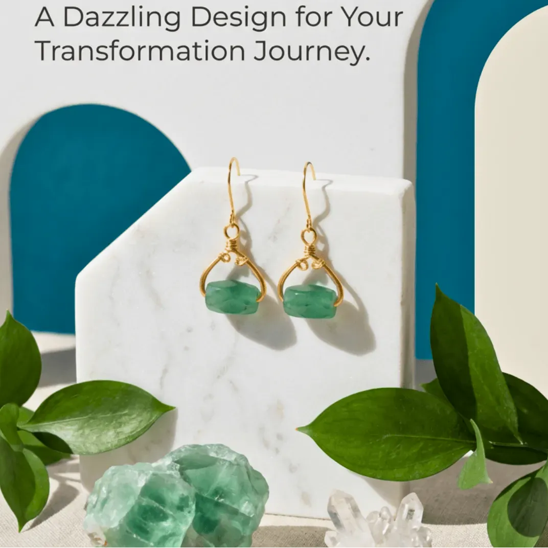 Gold earrings with green stones on a white marble stand with green leaves and crystals in the foreground.