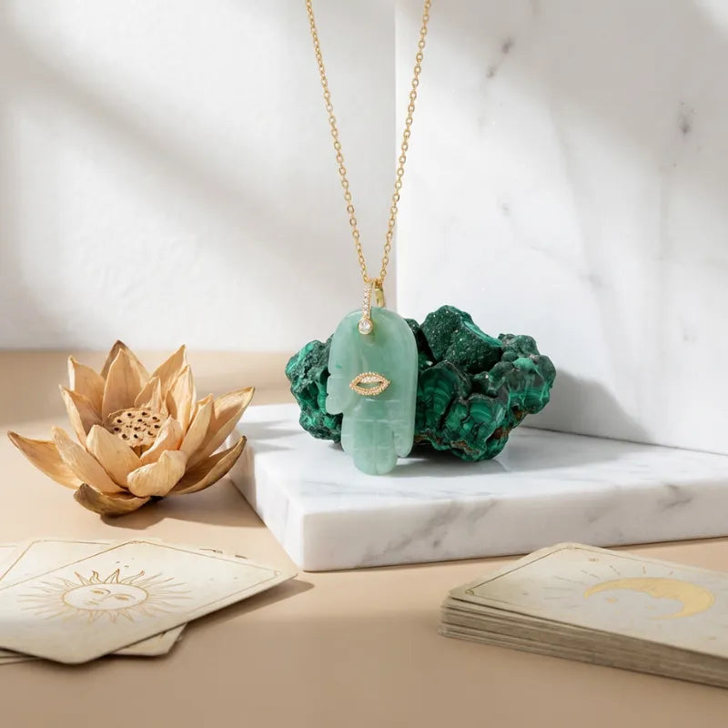 Green stone pendant on a gold chain with tarot cards and a wooden lotus flower on a marble surface.