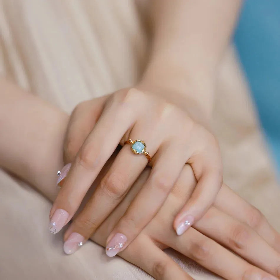 Close-up of a hand wearing a gold ring with a green gemstone, with a blurred background.