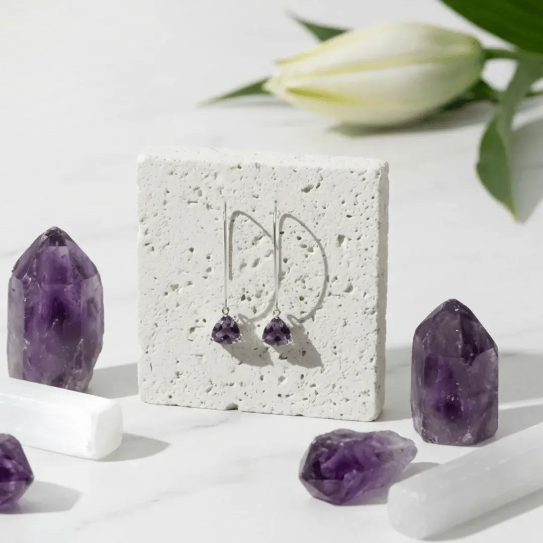 Silver earrings with purple stones on a white stone display card, surrounded by amethyst crystals.