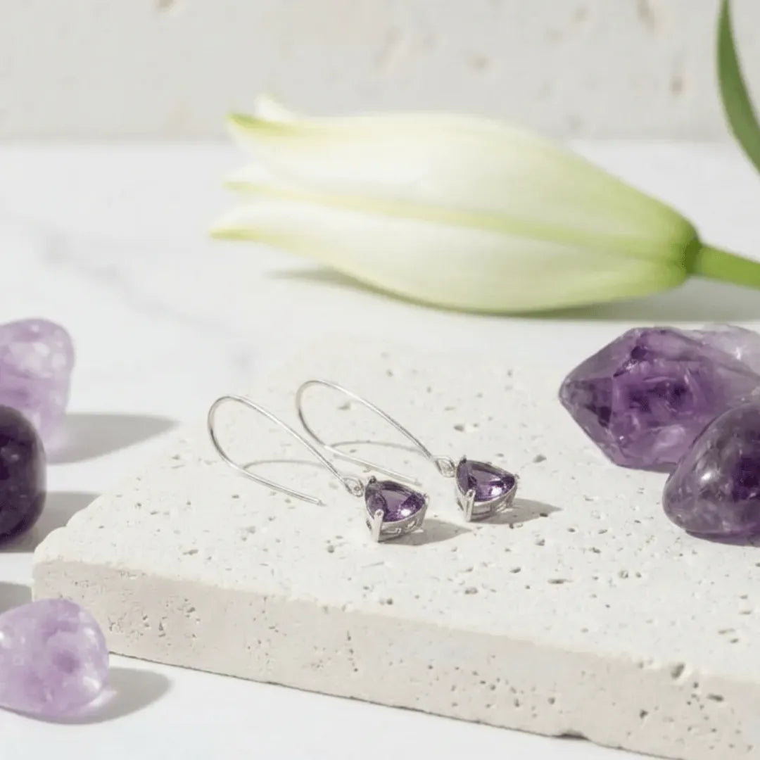 Heart-shaped purple gemstone earrings on a white stone surface with amethyst stones and a tulip.