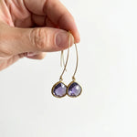 A hand holding a pair of gold hoop earrings with faceted purple teardrop gemstones against a plain light gray background.