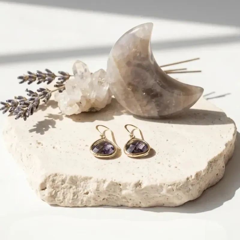 Gold earrings with purple stones on a stone surface with lavender and a stone crescent moon.