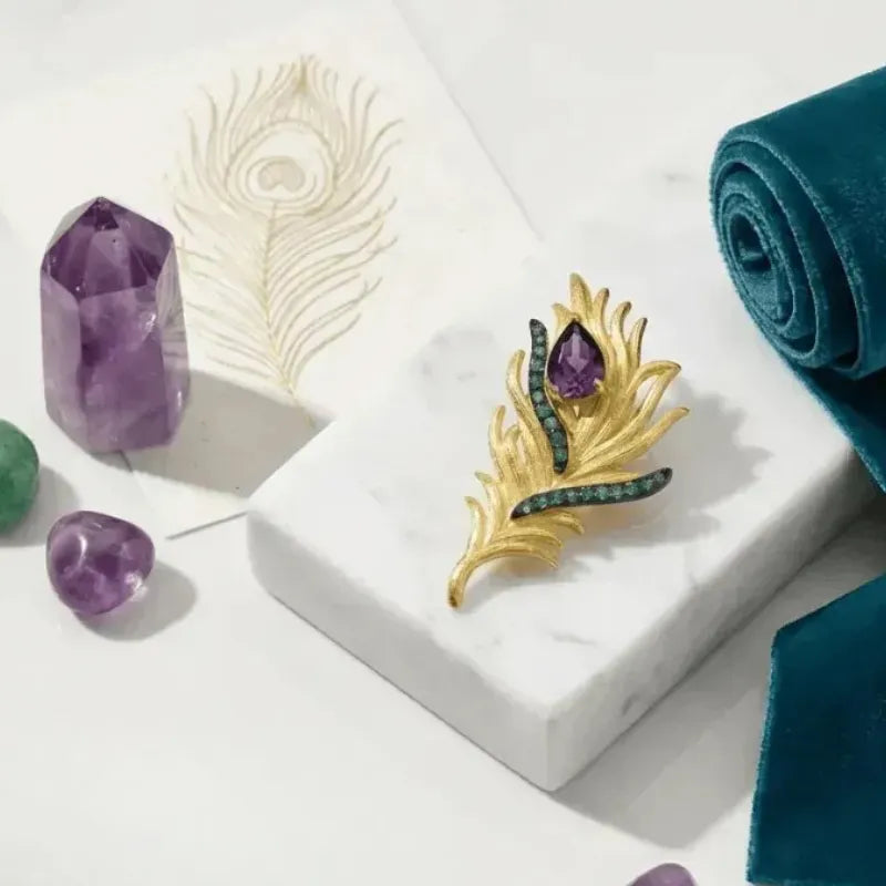Gold feather-shaped brooch with purple and green stones on a white surface with peacock feathers and crystals.