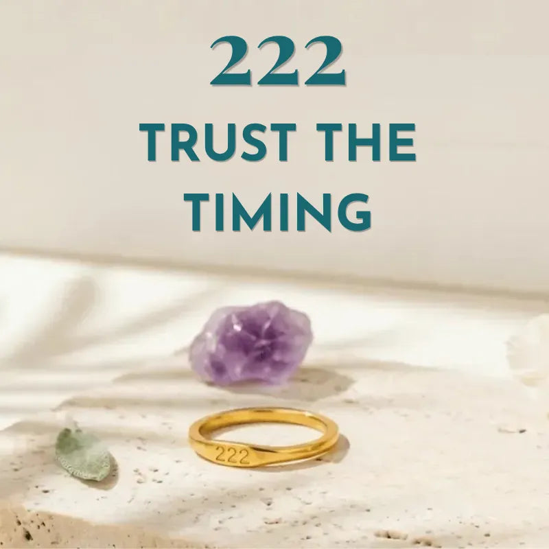 Gold ring with '222' on a stone surface with an amethyst crystal, text 'TRUST THE TIMING' above.