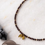 Close-up of the Garnet Moon Necklace with brown garnet beads and gold crescent moon pendant on a textured white surface, the original design as seen on The Vampire Diaries, handcrafted by Andrea of Mystic Soul Jewelry.