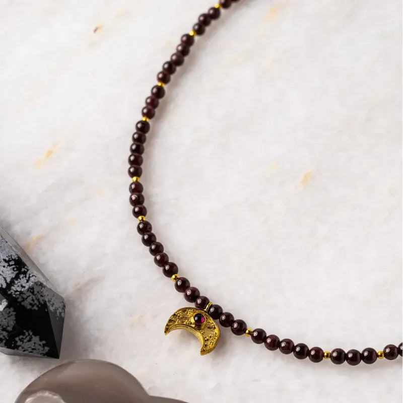 Close-up of the Garnet Moon Necklace with brown garnet beads and gold crescent moon pendant on a textured white surface, the original design as seen on The Vampire Diaries, handcrafted by Andrea of Mystic Soul Jewelry.