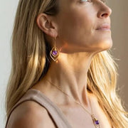 Close-up of a woman wearing gold earrings with a purple gemstone against a neutral background