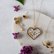 Gold jewelry set with heart-shaped pendant and bee earrings on a marble surface with purple flowers.