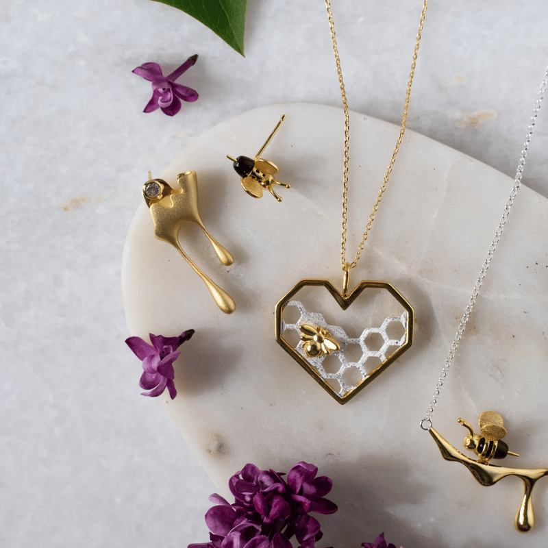 Gold jewelry set with heart-shaped pendant and bee earrings on a marble surface with purple flowers.