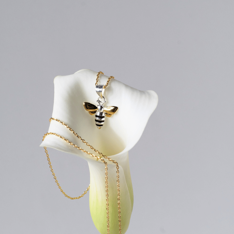 Gold necklace with a bee pendant on a white calla lily against a gray background