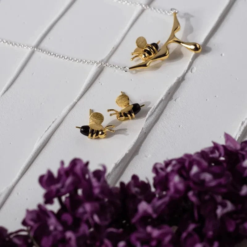 Gold bee-shaped earrings on a white surface with purple flowers.