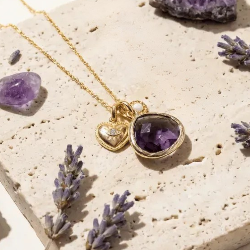 Gold necklace with an amethyst pendant on a textured surface with lavender flowers.