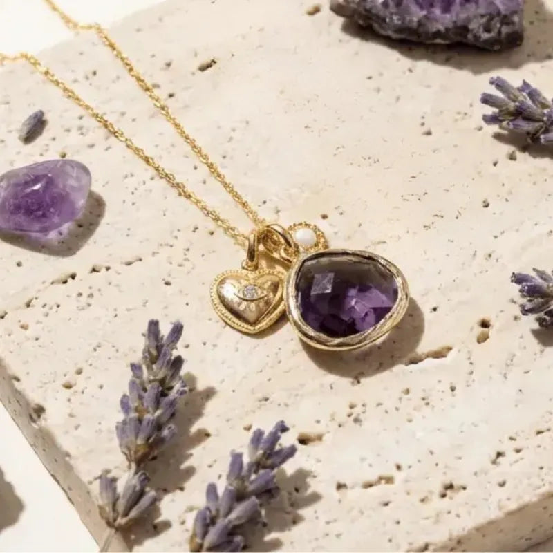 Gold necklace with an amethyst pendant on a textured surface with lavender flowers.