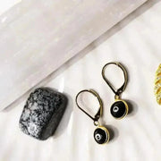 A pair of gold-plated black evil eye earrings displayed alongside a textured black stone and other small gold-tone jewelry items. 