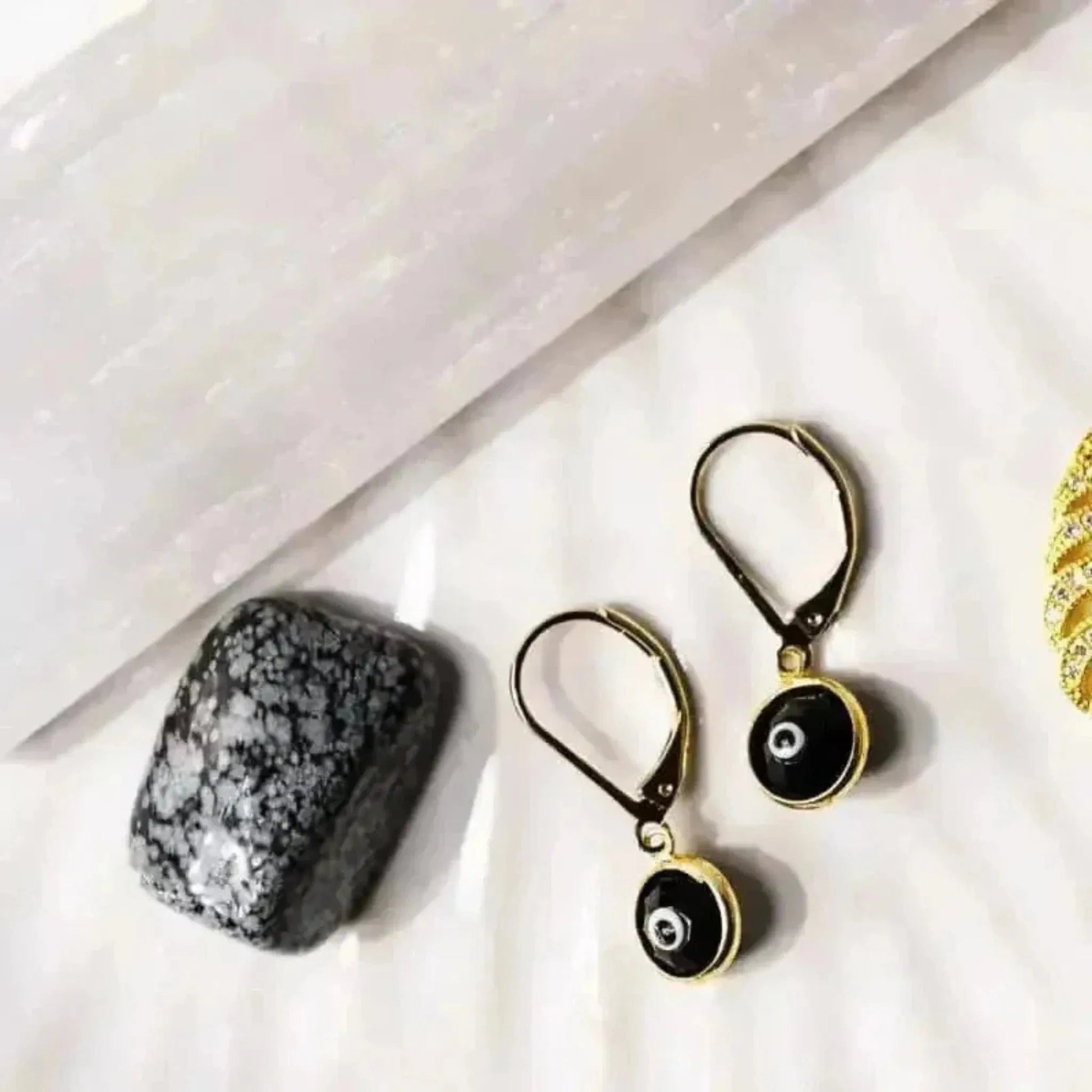 A pair of gold-plated black evil eye earrings displayed alongside a textured black stone and other small gold-tone jewelry items. 