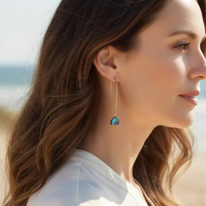 Woman wearing capri blue Cleo drop earring in gold on beach, ocean-tone jewelry