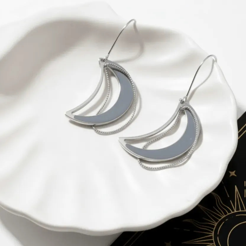 Two moon-shaped necklaces, one gold and one silver, on a white plate with a black card featuring star and moon designs.