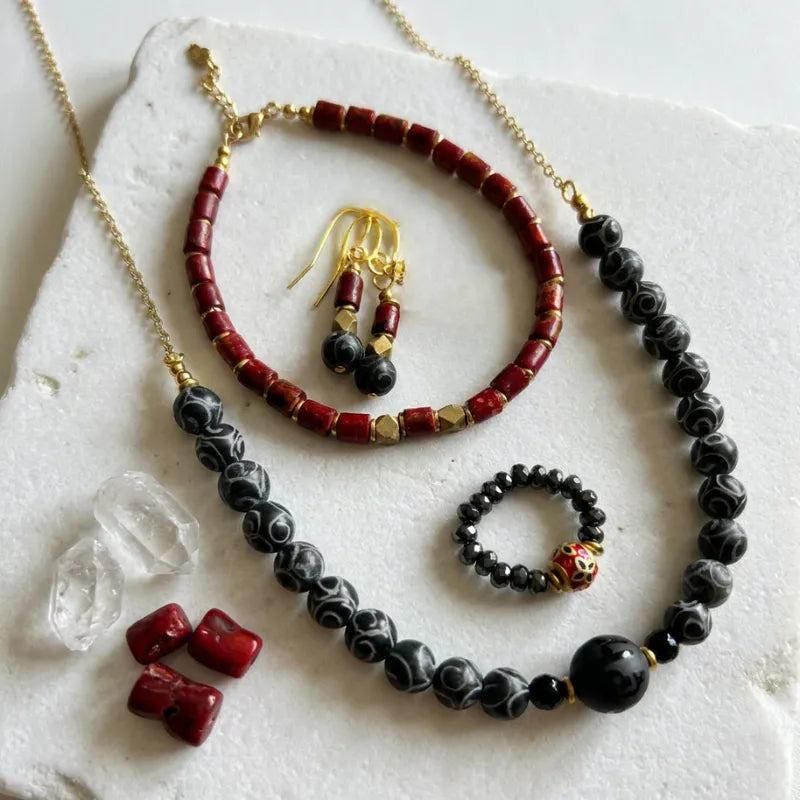 Agate dzi bead necklace with red coral bracelet, brass drop earrings and pyrite lucky bead ring on white stone surface