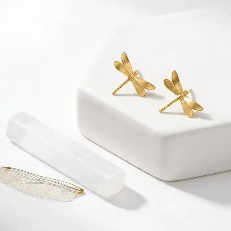 Gold dragonfly earrings on a white surface with a white box.