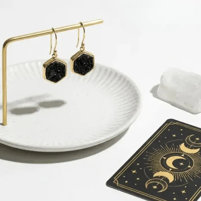 Black druzy earrings on a gold stand with a white dish and tarot card in the background.