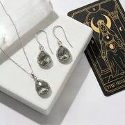 Silver jewelry set with teardrop-shaped stones on a white surface next to a tarot card.
