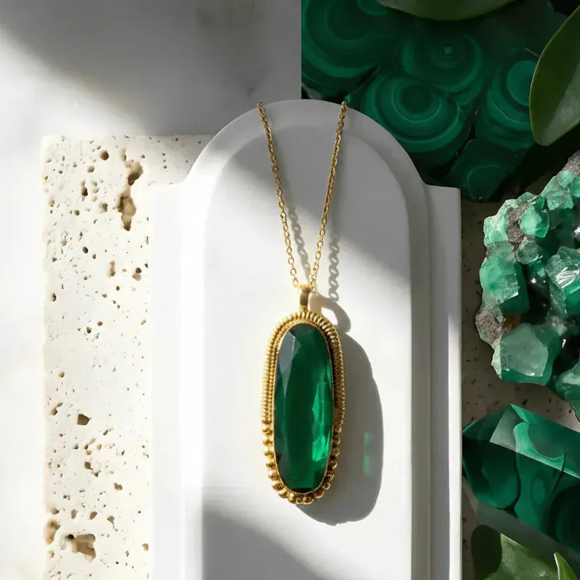 Gold necklace with a green pendant on a white surface with green crystals and leaves in the background.