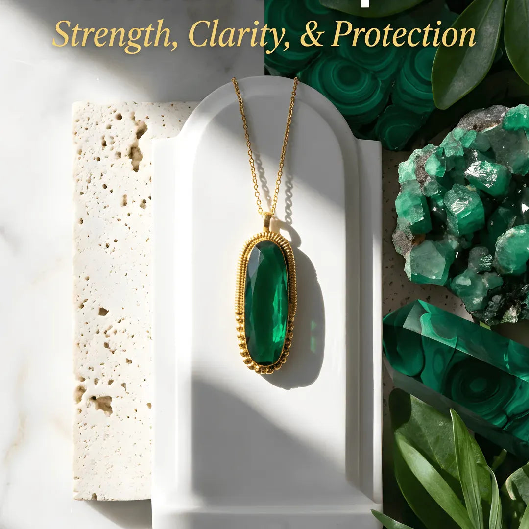 Gold necklace with a green pendant on a white surface with green crystals and leaves in the background.