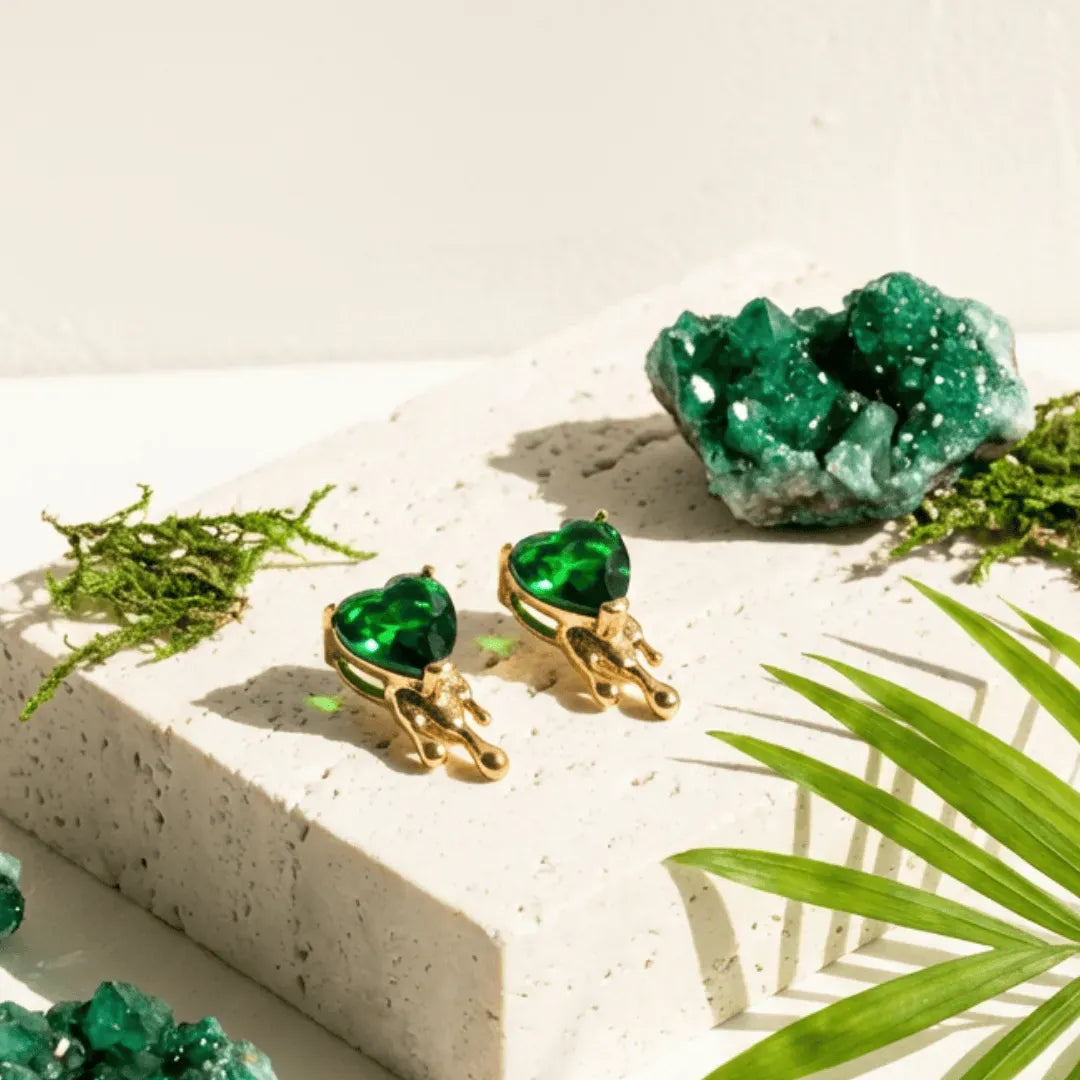 Green heart-shaped earrings with gold accents on a light surface with greenery.