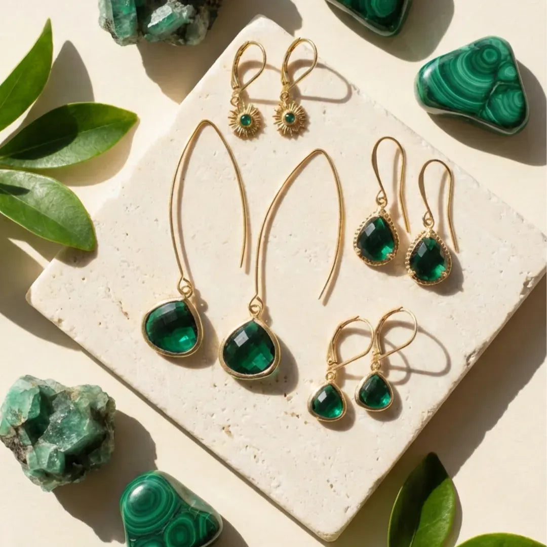 Set of green gemstone earrings with gold frames on a stone surface surrounded by green stones and leaves.