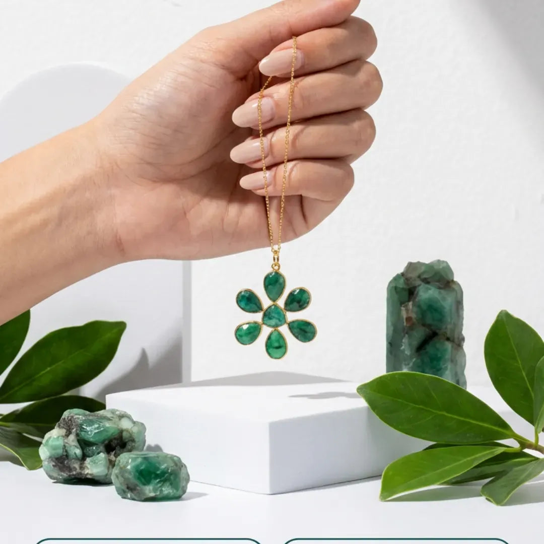 Hand holding a gold necklace with a green floral pendant, surrounded by green stones and leaves on a white background