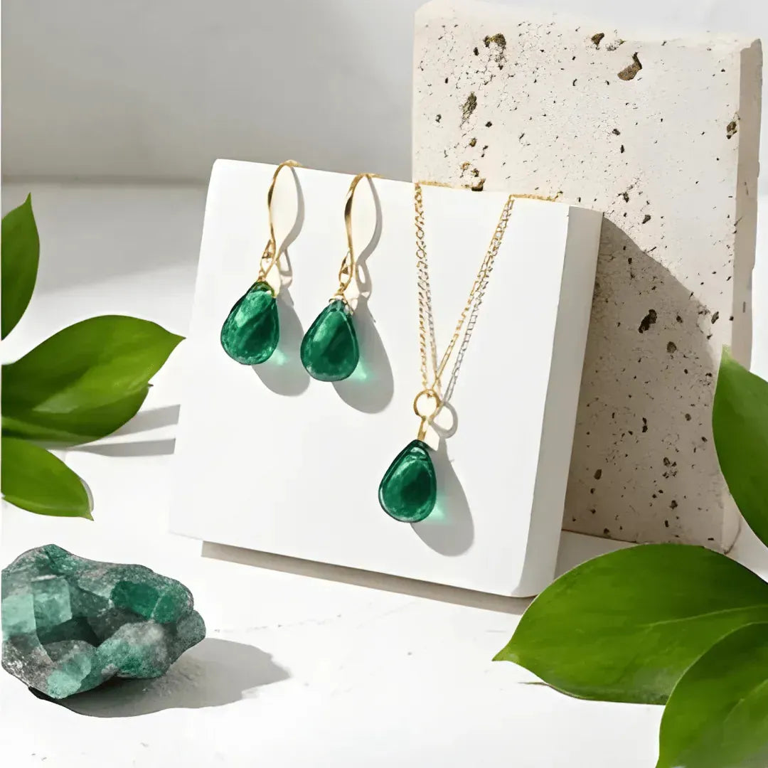 Green teardrop jewelry set with gold accents on a white stand, surrounded by green leaves.