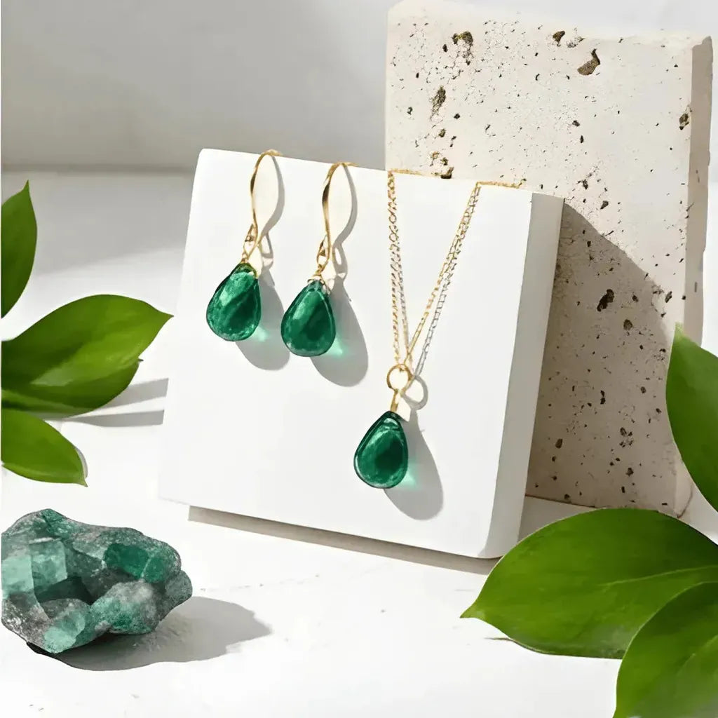 Green teardrop jewelry set with gold accents on a white stand, surrounded by green leaves.