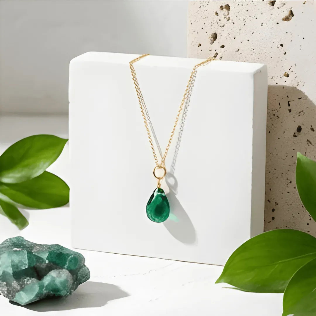 Gold necklace with a green teardrop pendant on a white stand, surrounded by green leaves and crystals.