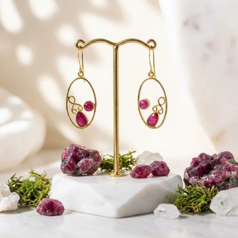 Gold earrings with pink gemstones on a marble stand with natural elements.