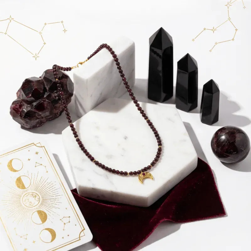 Necklace with maroon beads and gold accents on a marble stand, surrounded by crystals and tarot cards.