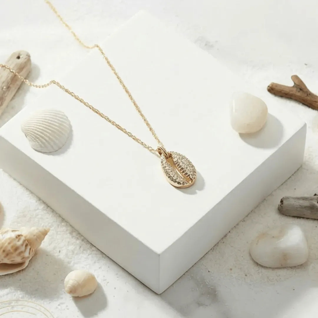 Gold necklace with a shell pendant on a white surface surrounded by seashells and driftwood.