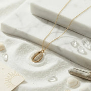 Gold necklace with a shell pendant on a sandy surface with shells and driftwood.