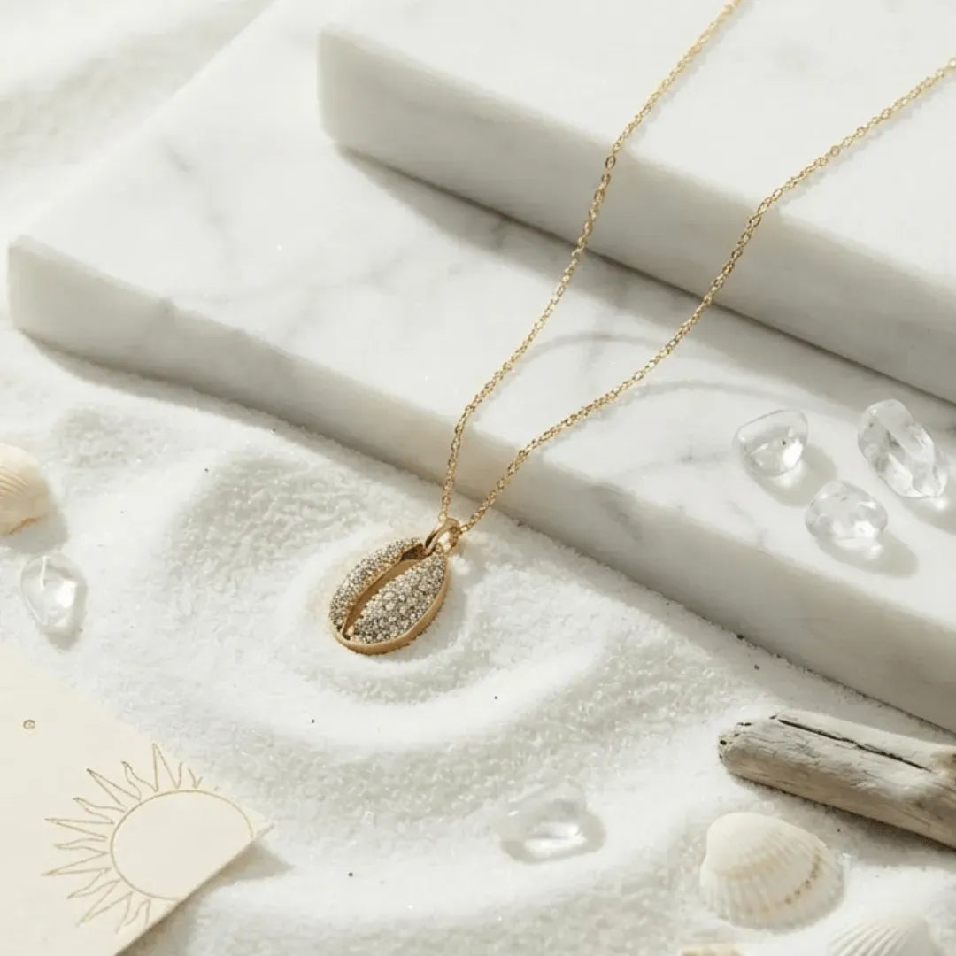 Gold necklace with a shell pendant on a sandy surface with shells and driftwood.