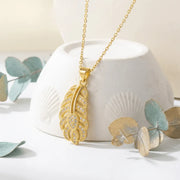 Gold necklace with a leaf-shaped pendant on a white surface with eucalyptus leaves.